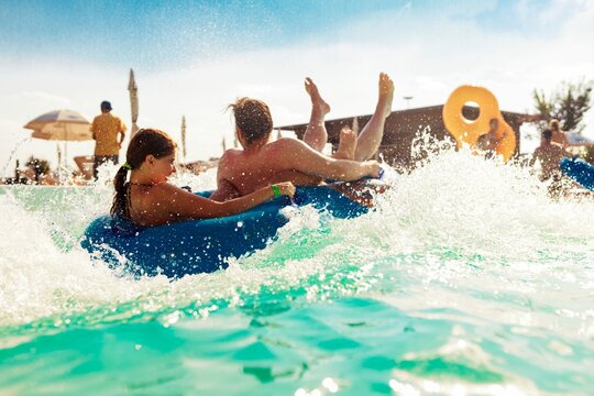 Two friends, a woman and a man, are shown enjoying a thrilling aqua ride in a water park on a bright summer day, getting splashed as they plunge into the turquoise water