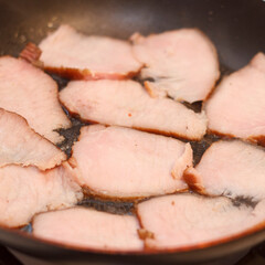 Slices of meat cooking in a frying pan. Meat is light pink with a crispy edge, arranged evenly in pan