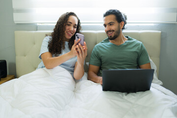 Happy couple relaxing in bed, using laptop and smartphone
