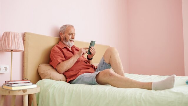 Senior man relaxing on bed using smartphone in modern apartment