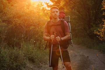 Handsome man hiker with backpack and poles walking through forest with warm sunset.