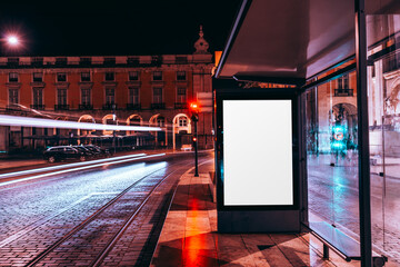 Blank illuminated billboard mockup at a bus stop in Lisbon at night, with long exposure light trails of passing vehicles and reflections on wet cobblestone street creating a dynamic urban scene