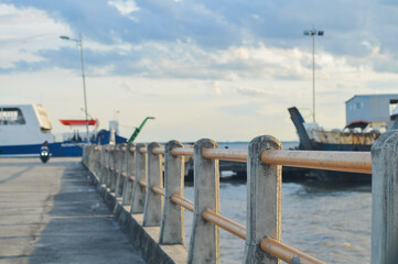 Concrete pier with safety railing at a seaport. Blurry view of a ferry ship and sea water in the background under cloudy sky. Transportation, travel, and maritime industry concept.