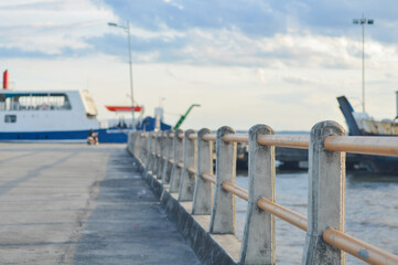 Naklejka premium Concrete pier with safety railing at a seaport. Blurry view of a ferry ship and sea water in the background under cloudy sky. Transportation, travel, and maritime industry concept.