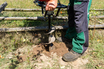Worker drilling hole for wooden post with handheld gasoline powered  auger drill in very rough, dry soil.