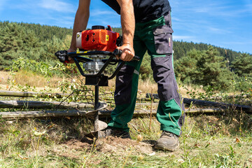 Worker drilling hole for wooden post with handheld gasoline powered  auger drill in very rough, dry soil.