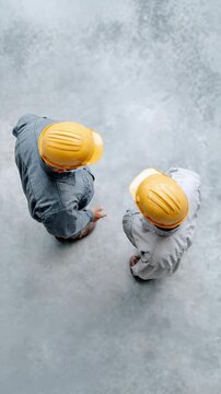 Two construction workers wearing hard hats are discussing a project on a concrete floor, captured from a top view perspective, emphasizing their collaboration and communication