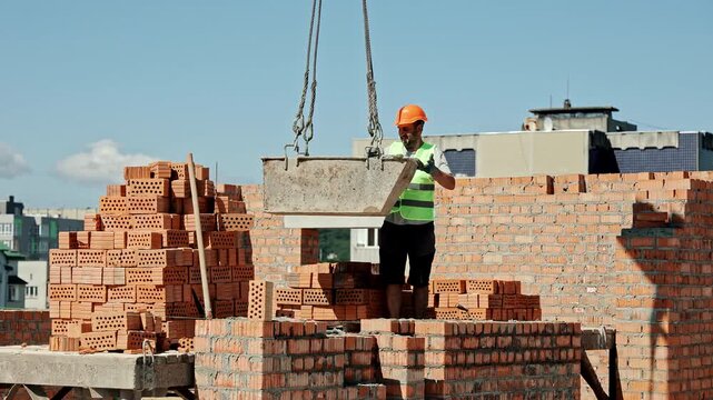 Construction worker in safety vest and helmet guiding suspended metal container at brick wall site, industrial scene with clear sky background and stacked red bricks