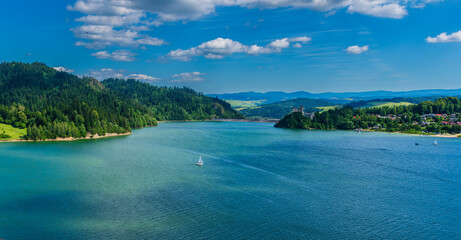 Turquoise waters of Czorsztyn Lake with sailboats gliding past the forested hills and medieval...