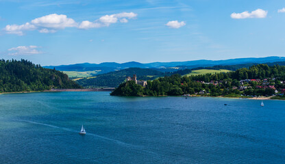 Historic Czorsztyn Castle perched on a hill above the calm waters of the lake, with a white sailing boat passing by. Perfect for travel brochures, postcards, cultural tourism, and advertising family 