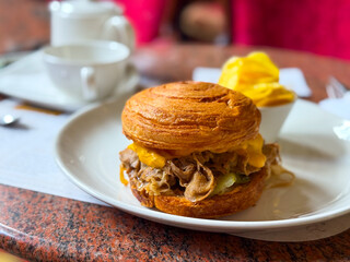 Croissant sandwich with melted cheese, meat, and vegetables served on white plate. Gastronomy, lifestyle, and modern dining representing fast casual cuisine, flavor, and contemporary eating.