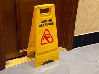 Yellow caution sign with wet floor warning placed indoors near wooden door. Safety, hygiene, and...
