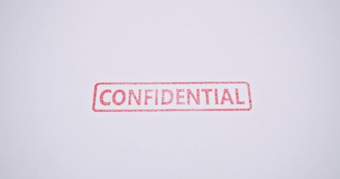 A close-up shot captures the moment a hand presses a red &ldquo;CONFIDENTIAL&rdquo; stamp onto a white sheet of paper. This real-life footage underscores the importance of safeguarding sensitive information.