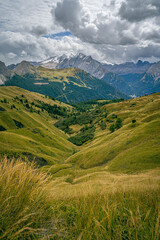 Obraz premium Green alpine hills in the Dolomites leading toward towering cliffs and distant mountain range beneath dramatic cloudy sky panoramic view