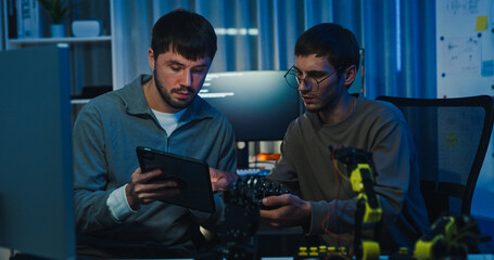 Team adult male engineer collaborate on robotic hand project using AI code solutions machine learning at tech workspace night. Artificial intelligence interface, collaboration prototype development.
