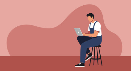 Young man in overalls working intently on a laptop while seated on a stool. Concept of remote work, online learning, or modern tech use. Copy space.