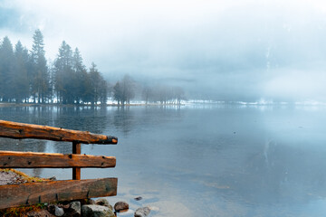 A calm lake is shrouded in thick fog, creating an ethereal setting. Trees line the shore, adding to the tranquil beauty of the quiet landscape during early morning hours.
