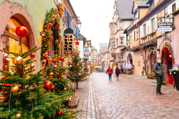 Colorful decorations and Christmas trees line the cobblestone streets as visitors explore the holiday market, enjoying the seasonal atmosphere on a cool day.
