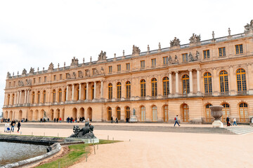 Visitors explore the grand exterior of Palatul Versailles, surrounded by manicured gardens and elegant sculptures, under a cloudy sky in France's historic site.