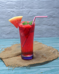 Refreshing summer drink with watermelon and ice in a glass, decorated with fruit slices, mint, and a straw, on a rustic blue wooden table with tropical details