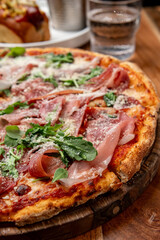 Tasty meat and greens pizza. A fresh pizza topped with cured meats and arugula served on a wooden board in a cozy setting.
