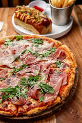 Pizza and hot dog on table. Tasty pizza topped with prosciutto and arugula served with a hot dog and fries, perfect for a casual dining experience.