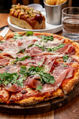 Tasty pizza with prosciutto and greens. A freshly baked pizza with prosciutto, arugula, and cheese sits on a wooden table next to fries and a hot dog.