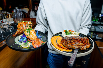 Lobster and steak served in a restaurant. A server holds two plates, one with grilled lobster and...