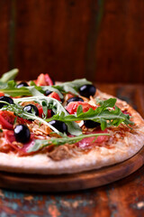 Pizza with salami, tomatoes and mozzarella cheese, black olives and arugula. Rustic wooden background. Soft focus. Copy space.	