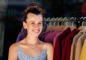 Smiling caucasian young female at outdoor clothing market displaying colorful sweaters