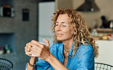 Portrait of a happy senior elderly woman holding a cup of coffee standing by window and relaxing at home