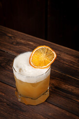 Citrus yellow cocktail with airy foam and dried orange garnish served in short glass on rustic wooden table