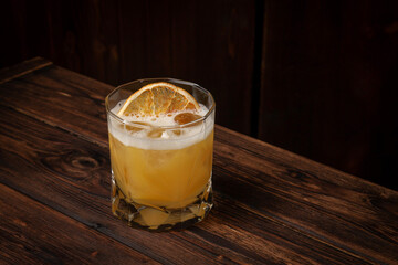 Classic whiskey sour cocktail with ice cubes and dried orange slice served in lowball glass on rustic wooden table
