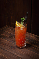Classic tequila sunrise cocktail with lemon wedge, rosemary sprig and ice cubes in tall crystal glass on rustic wooden table