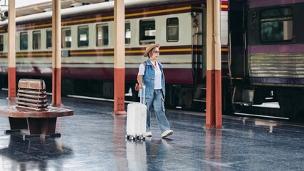 Older Asian woman traveler walking train station platform with white luggage for solo journey vacation