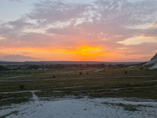 A serene landscape at sunset with vibrant orange and pink hues in the sky. The foreground features a grassy field with white patches, ideal for copy space.