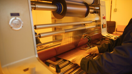 Gloved factory worker operating an industrial lamination machine, processing copper foil for...