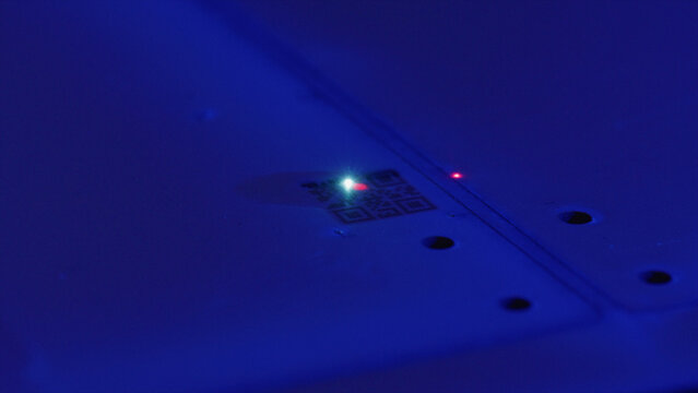 Laser engraving machine is engraving qr code on a metal surface, generating a bright green light and a red aiming beam in a dark blue environment - Powered by Adobe