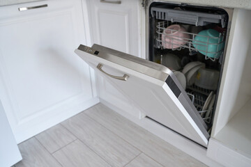 Open dishwasher with clean dishes in modern kitchen featuring wooden floor and white cabinets.