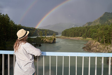 Female viewing double rainbow over scenic river and forest landscape in mountainous region.