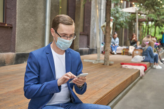 Young caucasian male in blue blazer using smartphone outdoors with face mask in urban setting.