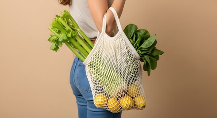 Woman in jeans holding a reusable mesh grocery bag filled with fresh produce like celery, lemons, and leafy greens.