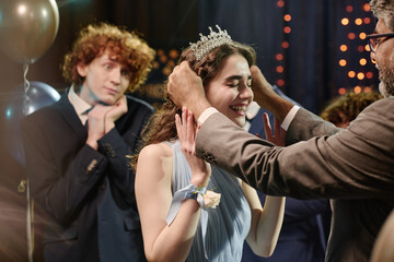 Caucasian teenage girl smiling while adult man placing tiara on her head during celebration, teenage boy with curly hair standing in background holding hands to face and watching