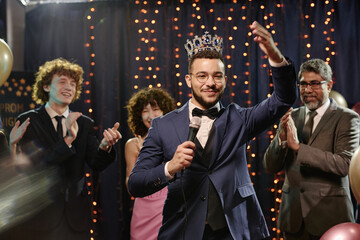 Teenage boy wearing crown holding microphone smiling and waving while standing on stage at prom...