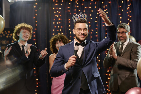 Teenage boy wearing crown holding microphone smiling and waving while standing on stage at prom event with teenagers and man clapping in background