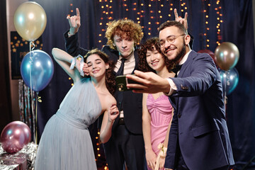 Group of multiethnic teenagers posing for selfie at party, smiling and making hand gestures, standing together in front of balloons and festive decorations, enjoying celebration