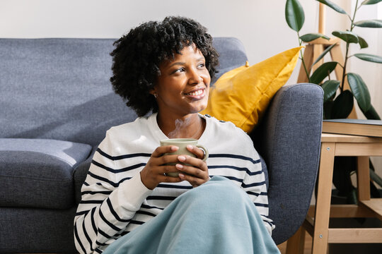 Young woman is sitting on the floor by the couch, wrapped in a blanket and holding a hot tea or coffee drink, enjoying a cozy winter day at home