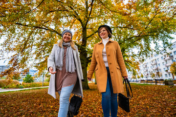 Two beautiful mature women walking in city park on autumn day. Front view