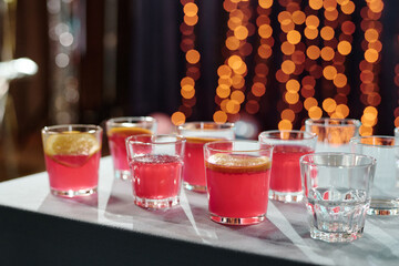 Multiple glasses with red beverages garnished with lemon slices standing on table, empty glass included, festive bokeh lights glowing in background, no people visible