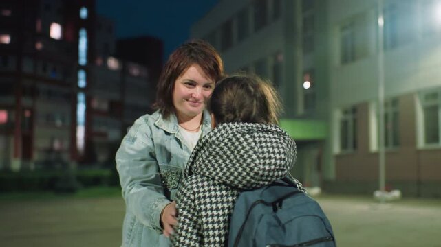 Woman lovingly looks into eyes of young girl wearing backpack and houndstooth coat outdoors near residential and school buildings at night, capturing warm emotional connection and quiet moment